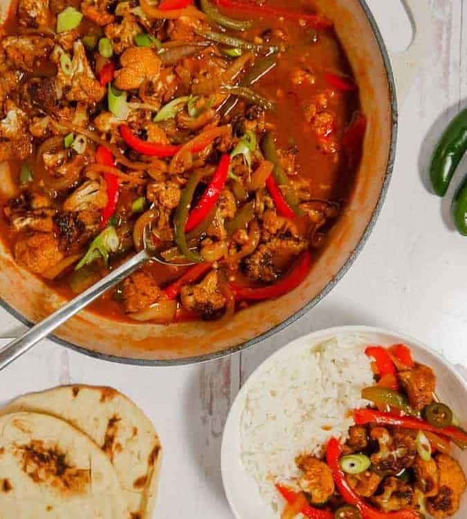 overhead photo of a pot filled with cauliflower, gravy, and bell peppers, with naan bread, bowl of gobi machurian and peppers on flatlay