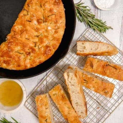 overhead photo of focaccia bread in a cast iron skillet, sliced bread, olive oil and rosemary
