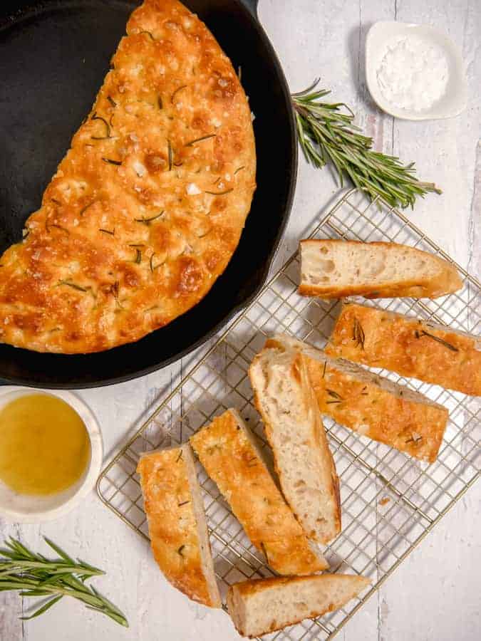 overhead photo of focaccia bread in a cast iron skillet, sliced bread, olive oil and rosemary