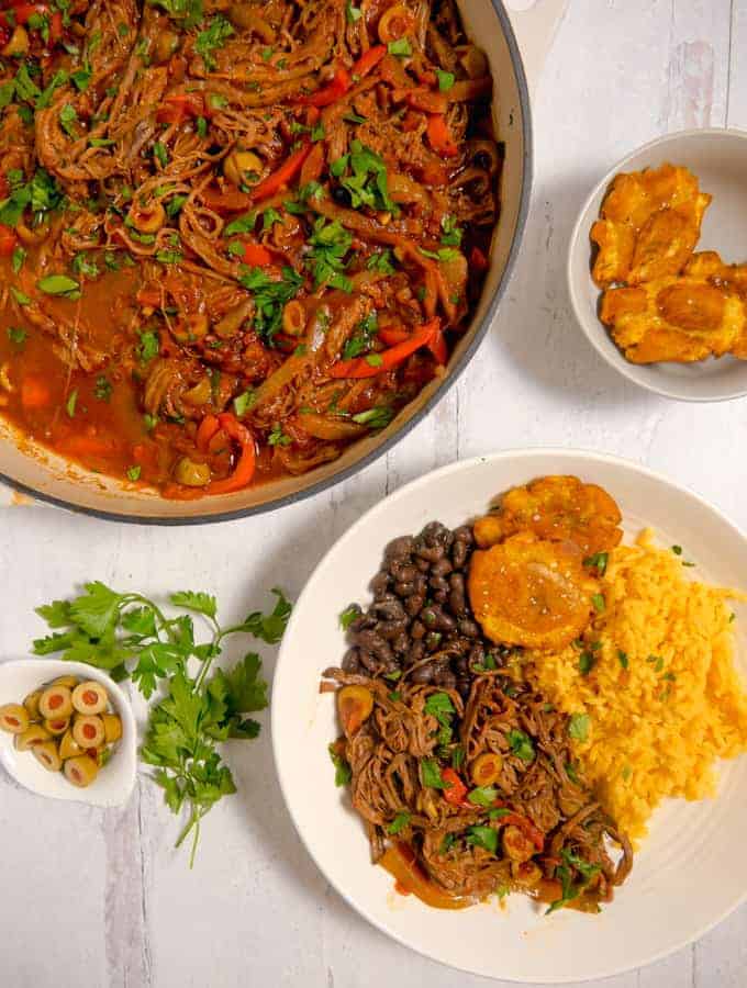 overhead photo of shredded beef in a pot with a plate of ropa vieja, yellow rice, black beans, and tostones