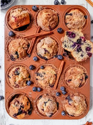 overhead photo of blueberry muffins in a muffin tin surrounded by fresh blueberries and cinnamon sticks