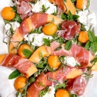 close up of melon wrapped in prosciutto on a plate with herbs and burrata cheese