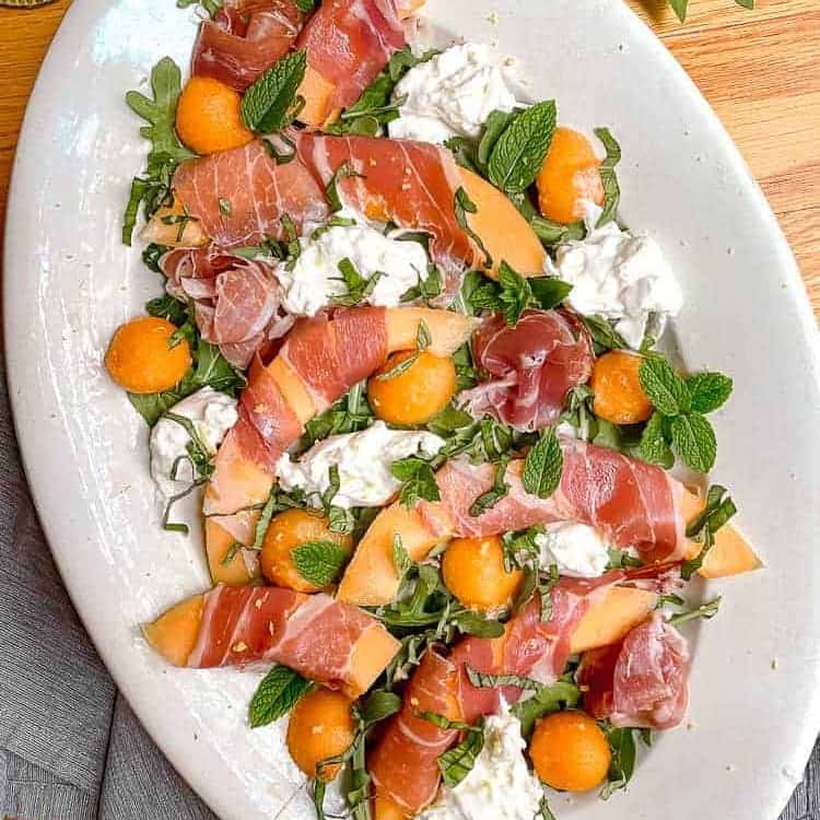 white serving dish with melon wrapped in prosciutto, fresh basil, mint, and burrata cheese