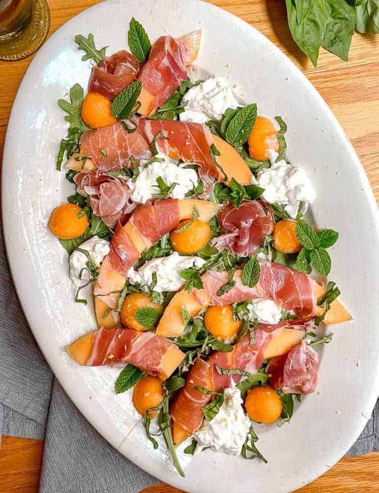 white serving dish with melon wrapped in prosciutto, fresh basil, mint, and burrata cheese