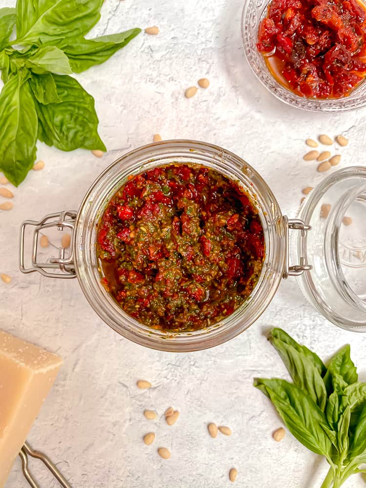 overhead photo of a glass jar filled with pesto and sun dried tomatoes, surrounded by fresh basil leaves and a cheese block