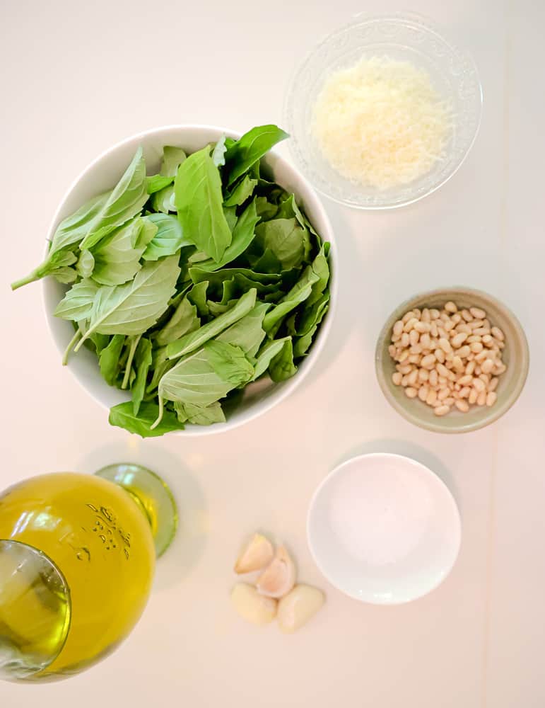 pesto ingredients on the counter: bowl of basil leaves, grated parmesan cheese, olive oil, garlic cloves, kosher salt and pine nuts