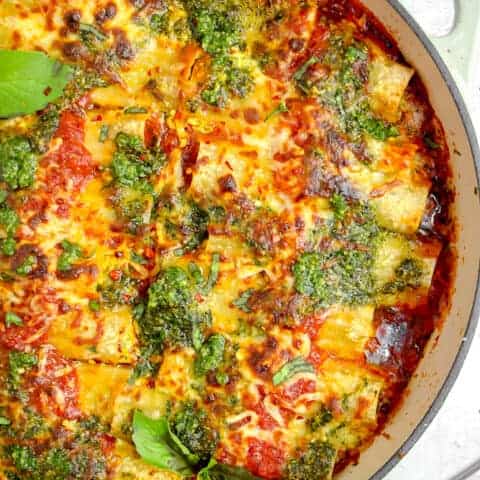 white casserole pan with red sauce, pesto stuffed manicotti, browned bubbly cheese and pesto on top with fresh basil leaves