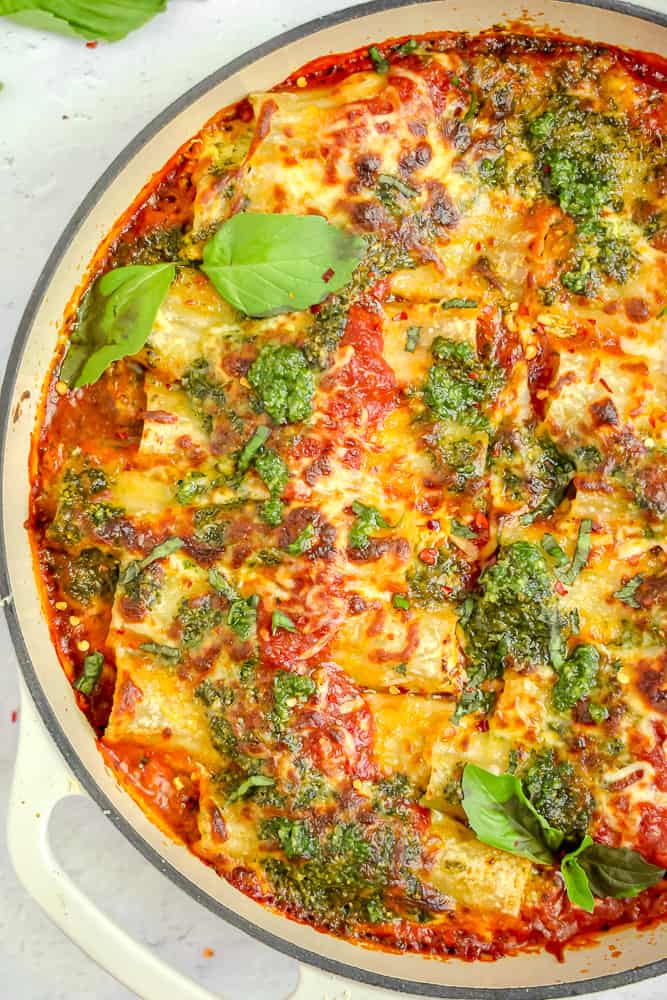 white casserole pan with red sauce, pesto stuffed manicotti, browned bubbly cheese and pesto on top with fresh basil leaves