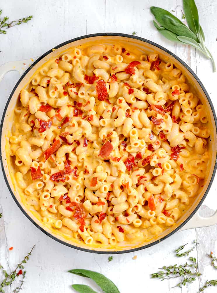 squash mac and cheese topped with crispy prosciutto in a skillet surrounded by herbs