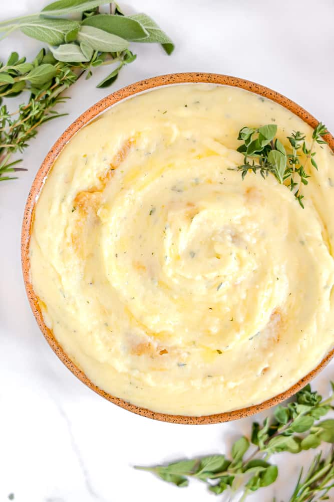 herbed mashed potatoes in a bowl with roasted garlic surrounded by herbs