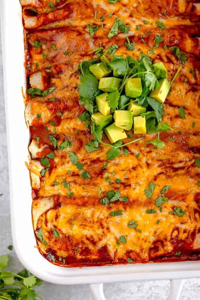 white baking dish with red enchiladas covered in melted cheese and topped with avocado and cilantro