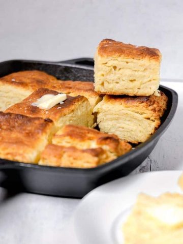 flakey buttermilk biscuits in a cast iron skillet stacked on each other and a slice of butter on top