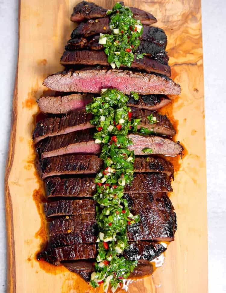 grilled flank steak sliced and topped with chimichurri over the top on a wooden block