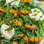 Arugula, grilled peaches, burrata, and basil dressing on a white plate on a cutting board