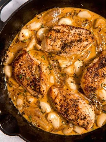 chicken surrounded by 30 cloves of garlic in a creamy sauce in cast iron skillet