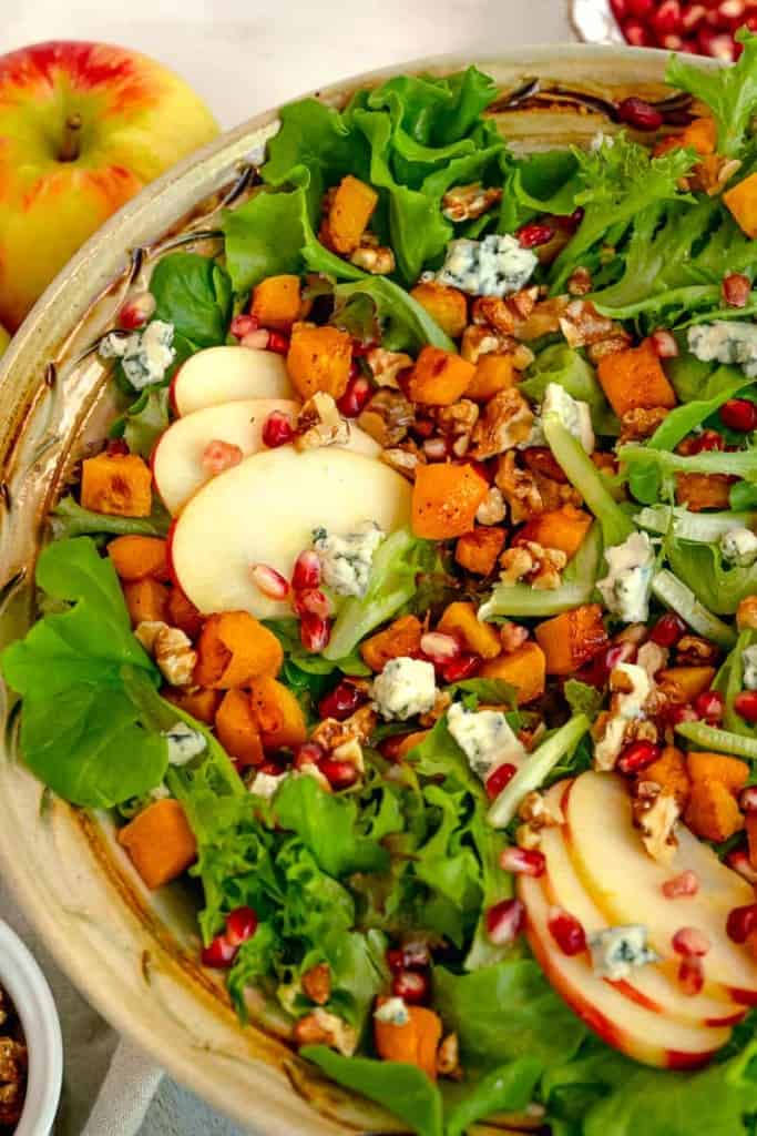 close up of mixed greens with sliced apples, butternut squash, pomegranate seeds, and walnuts on top