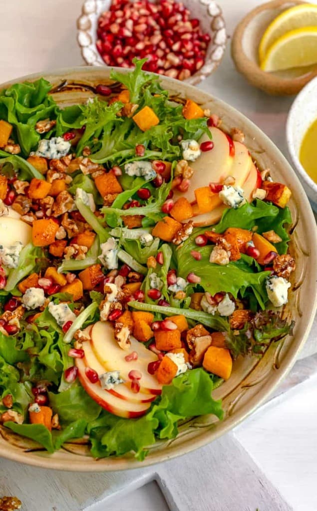 roasted butternut squash salad in a brown bowl with pomegranate seeds and lemon wedges in background