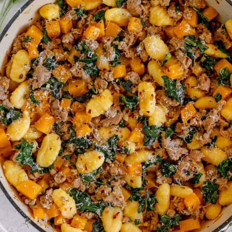 white skillet with gnocchi, butternut squash, kale, and hot italian sausage surrounded by fresh sage and thyme