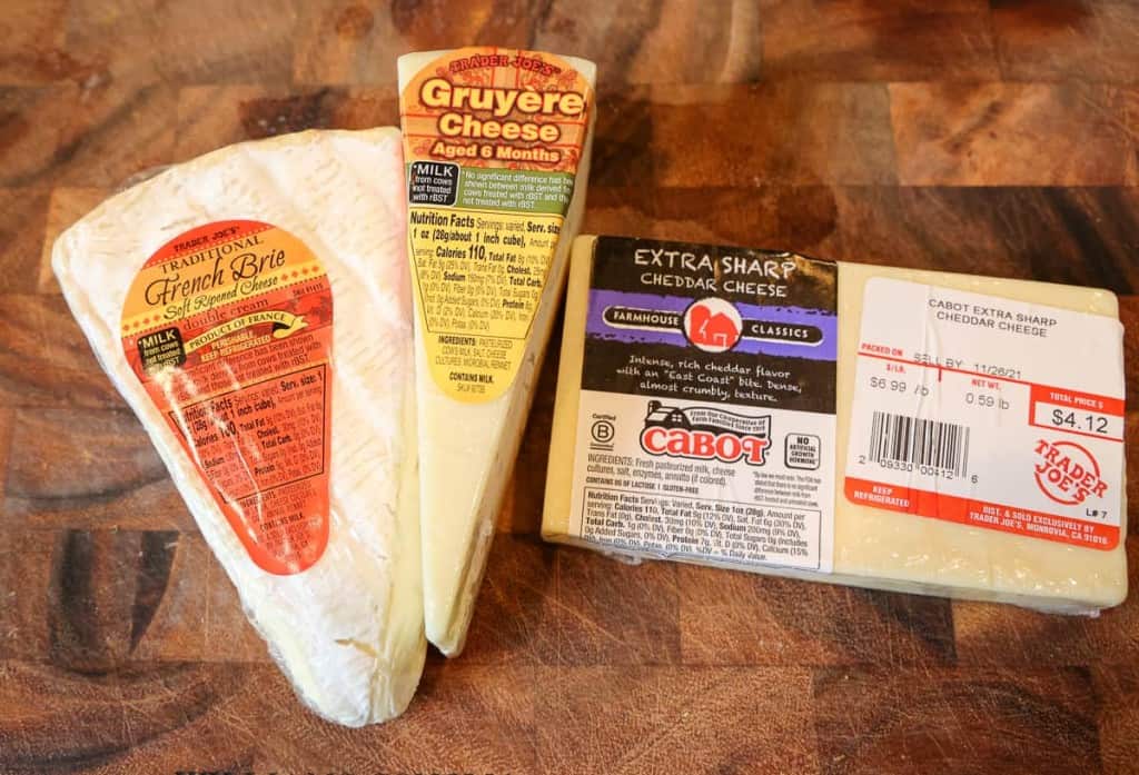gruyere, brie, and sharp cheddar cheeses in packaging on cutting board