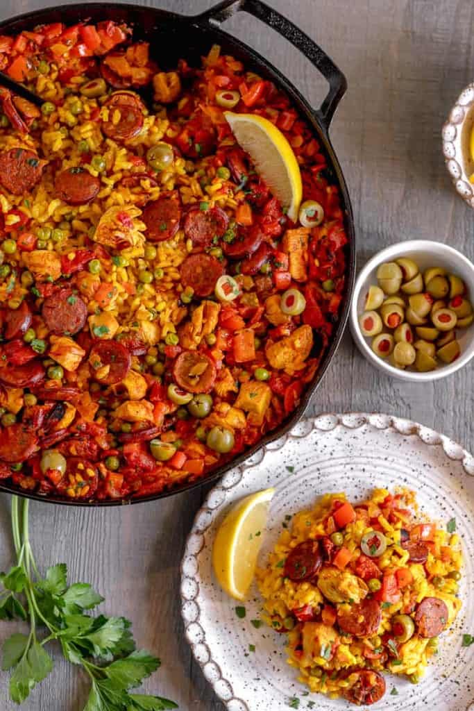 cast iron with chicken and chorizo paella with a white side plate with a serving of paella on top and lemon wedge on the side