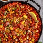 cast iron skillet filled with chicken and chorizo paella, topped with lemon wedges and surrounded by fresh parsley and green olives