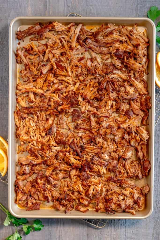 a gold baking sheet with shredded pork carnitas, surrounded by cilantro and orange slices