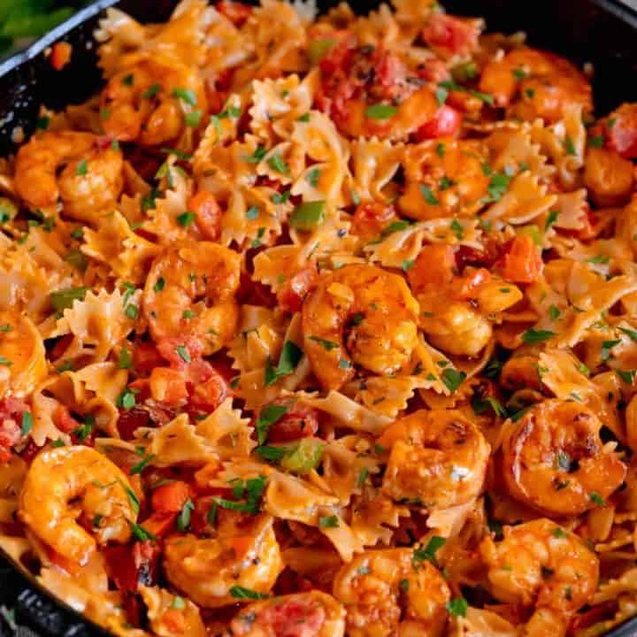 cast iron skillet filled with bowtie pasta, shrimp, topped with parsley and surrounded by fresh herbs on a grey background