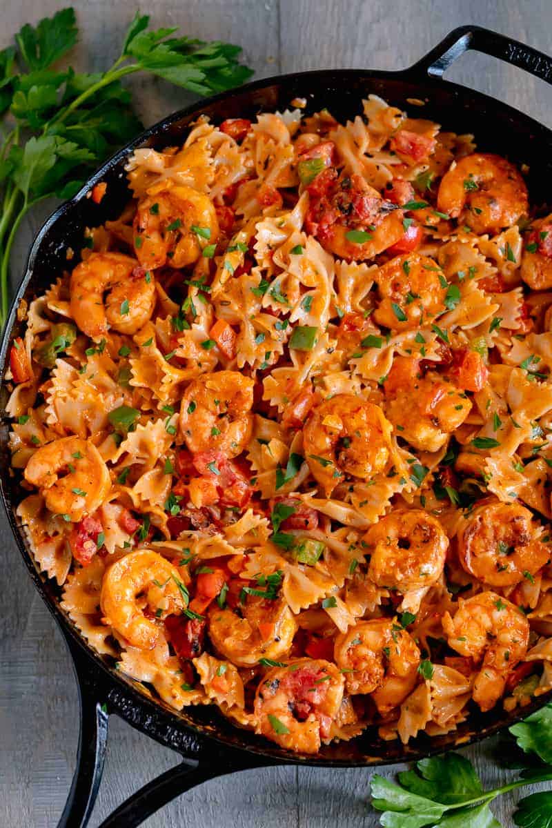 cast iron skillet filled with bowtie pasta, shrimp, topped with parsley and surrounded by fresh herbs on a grey background