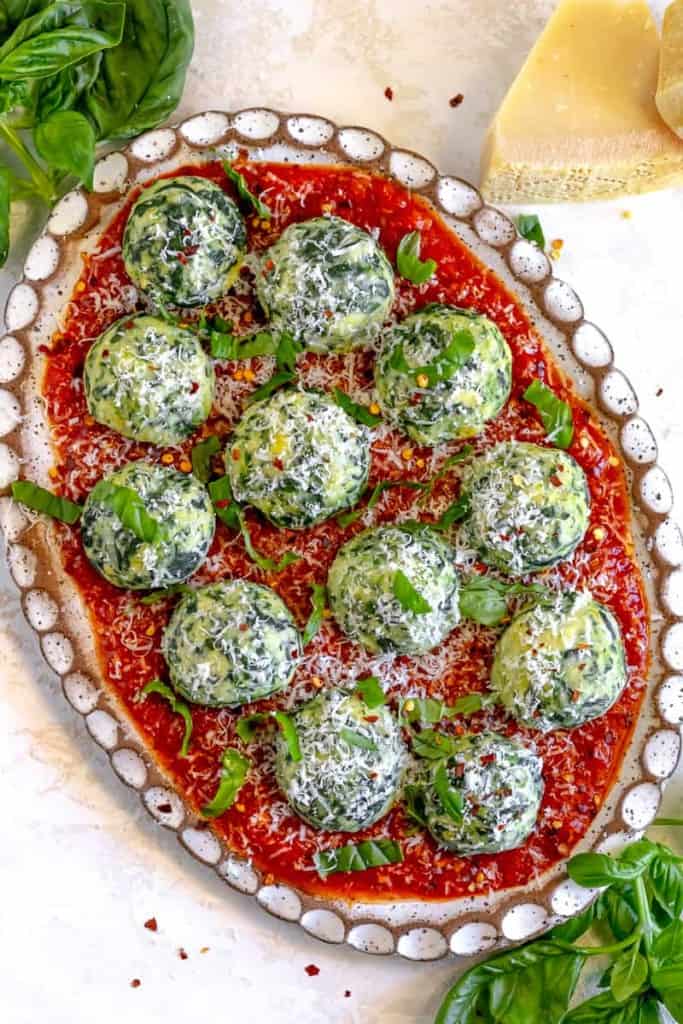 spinach ricotta gnudi over red sauce, topped with fresh parmesan, on an oval white and brown plate and surrounded by fresh basil