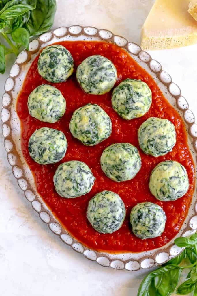 spinach and ricotta gnudi on a bed of red sauce on an oval plate surrounded by fresh basil and block of cheese