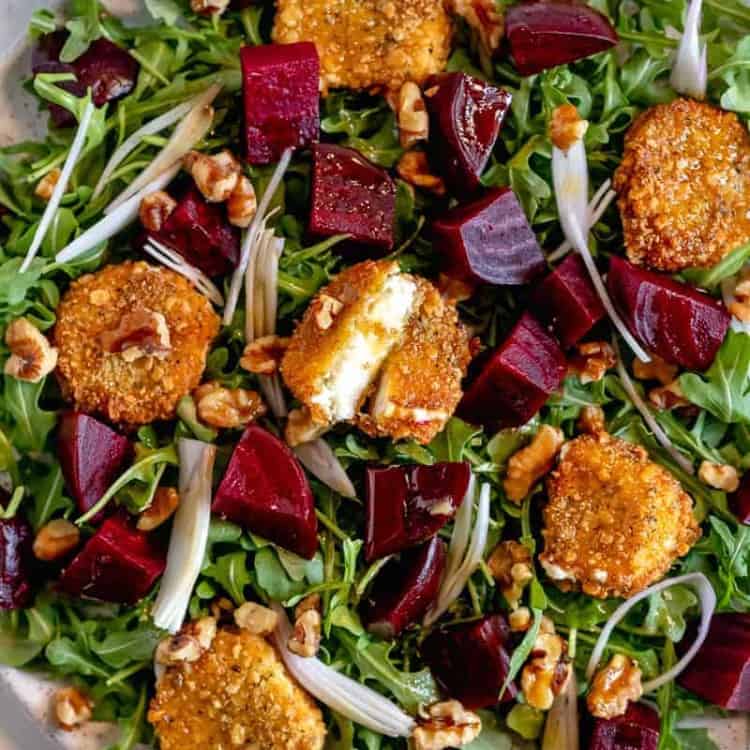close up of fried goat cheese cut in half surrounded by roasted beets on a bed or arugula on a white plate