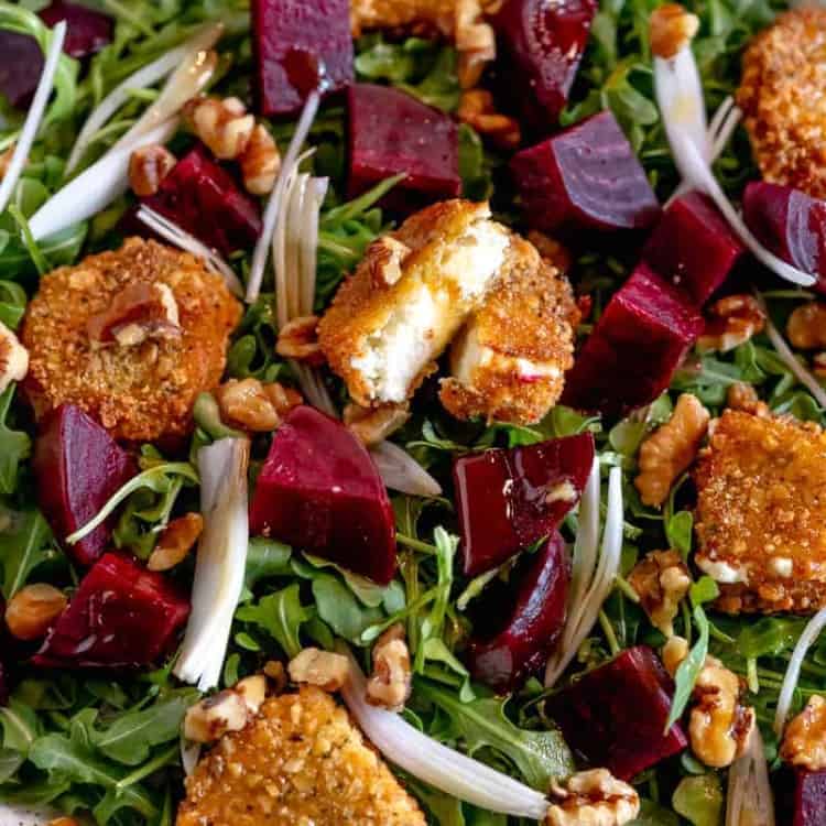 an arugula salad topped with fried goat cheese sliced down the middle, quartered roasted beets, and shallots on a white plate