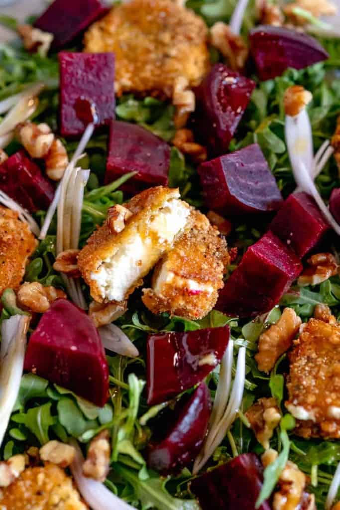 close up of fried goat cheese cut in half surrounded by roasted beets on a bed or arugula on a white plate