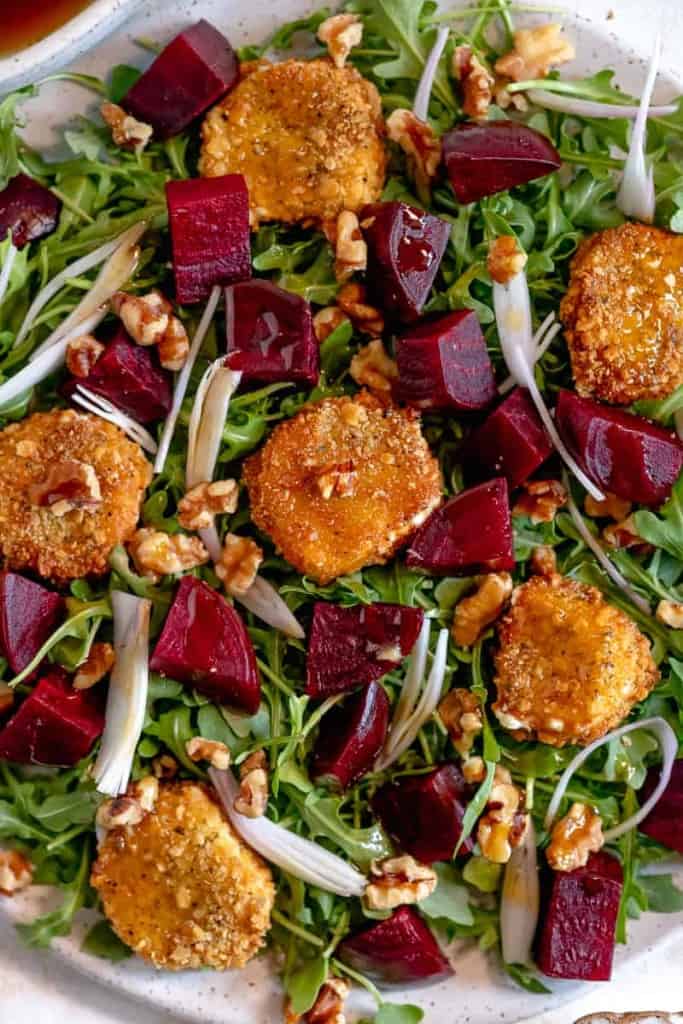 Arugula salad on a white plate topped with fried goat cheese, roasted beet slices, shallots, and walnuts.
