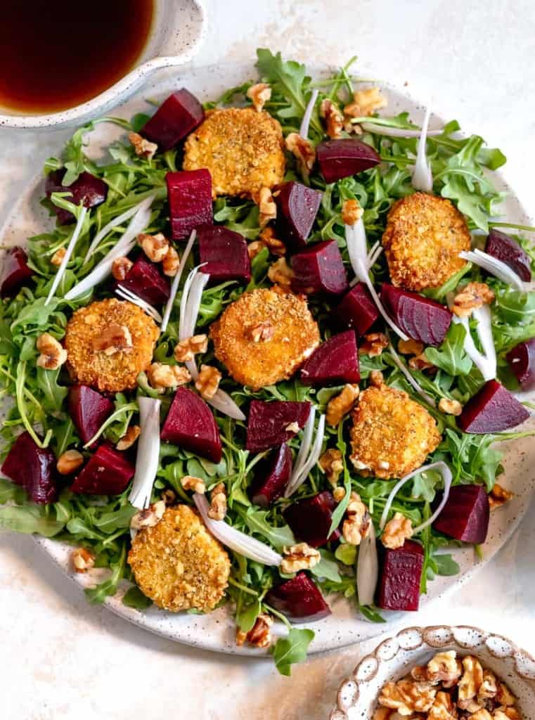 Arugula salad on a white plate topped with fried goat cheese, roasted beet slices, shallots, and walnuts.