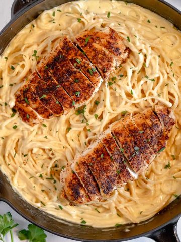 cast iron skillet on white background filled with creamy Alfredo pasta and sliced blacked chicken breast on top of pasta