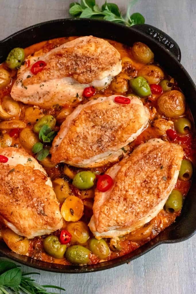 cast iron skillet with skin on chicken, surrounded by baby potatoes, castelvetrano olives, sliced red peppers, and fresh herbs on a grey background