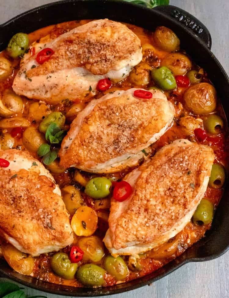 cast iron skillet with skin on chicken, surrounded by baby potatoes, castelvetrano olives, sliced red peppers, and fresh herbs on a grey background