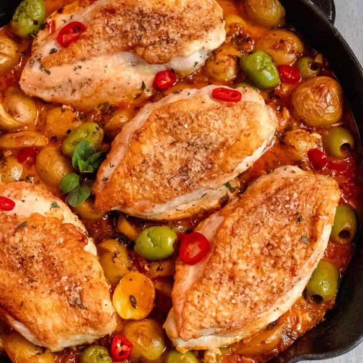 skin-on chicken breasts in a cast iron skillet surrounded by potatoes, red sauce, chile peppers, and green olives