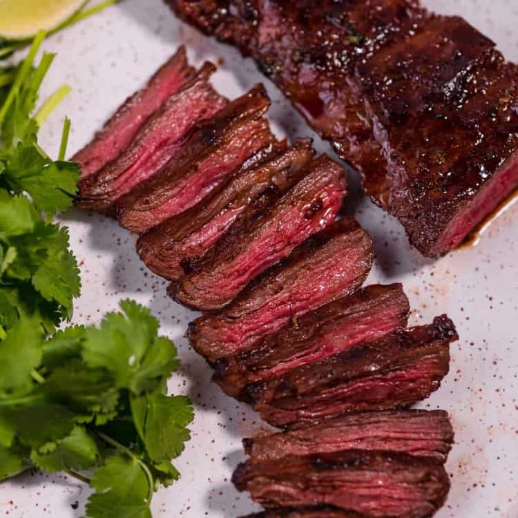 sliced skirt steak on a white plate next to fresh bunch of green cilantro