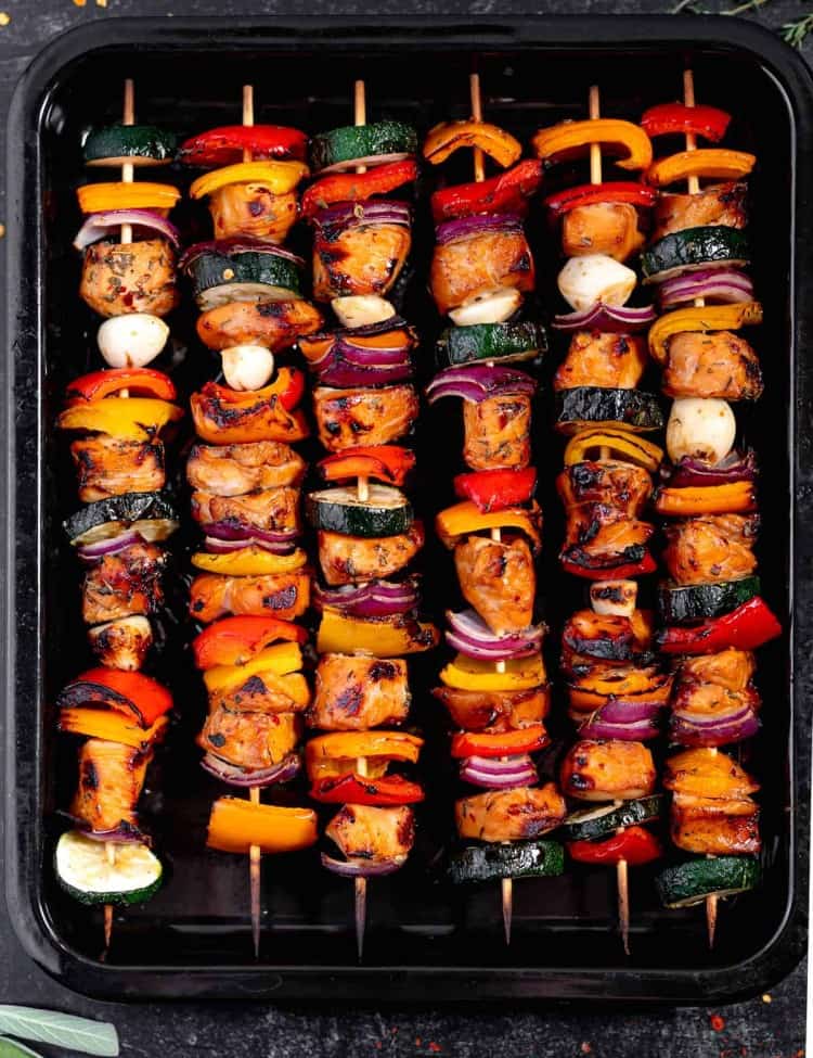 wooden skewers with a mix of chicken, bell peppers, red onions, garlic cloves, and zucchini, in a black pan on a black backdrop