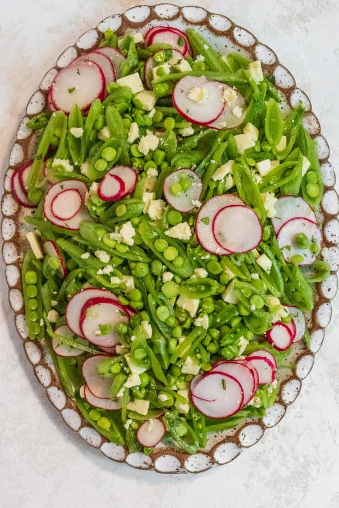 White oval and brown plate filled with sliced green snap peas and thinly sliced radishes on a marble background