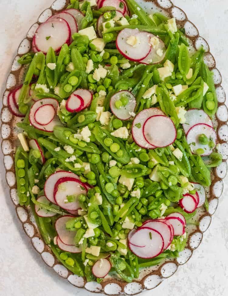 White oval and brown plate filled with sliced green snap peas and thinly sliced radishes on a marble background