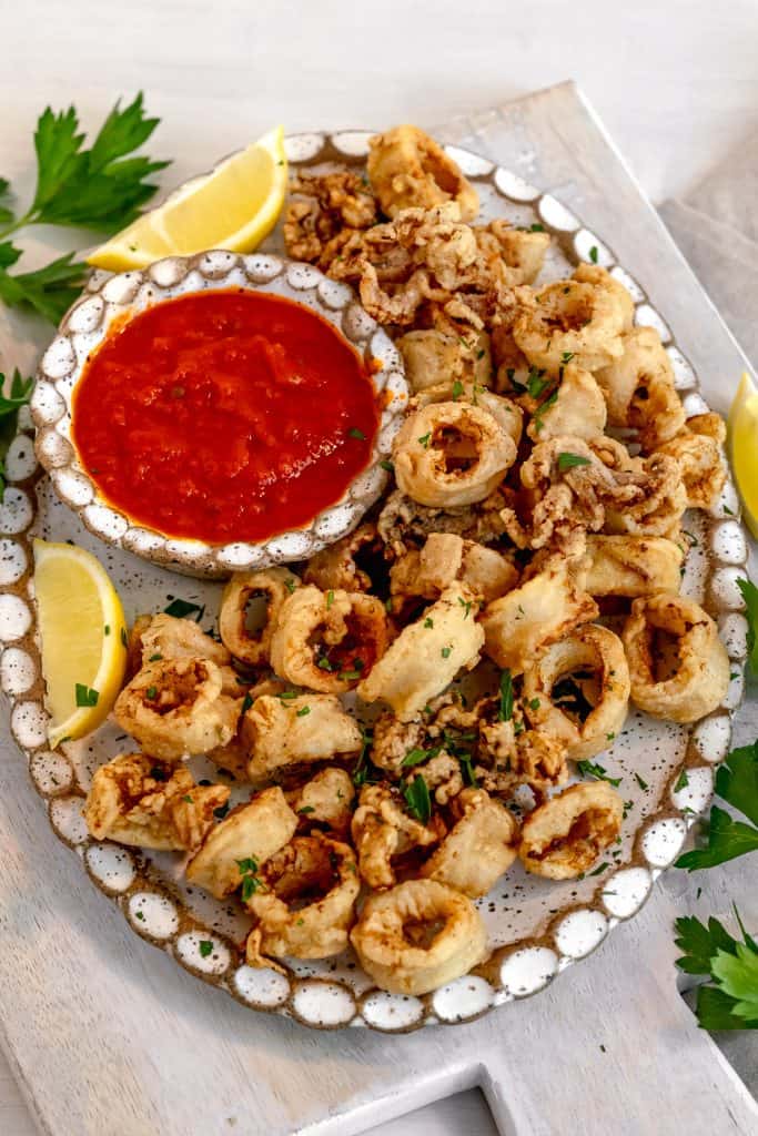 oval platter with crispy fried calamari and a cup of red sauce. Platter surrounded by lemon wedges and fresh parsley on a grey background