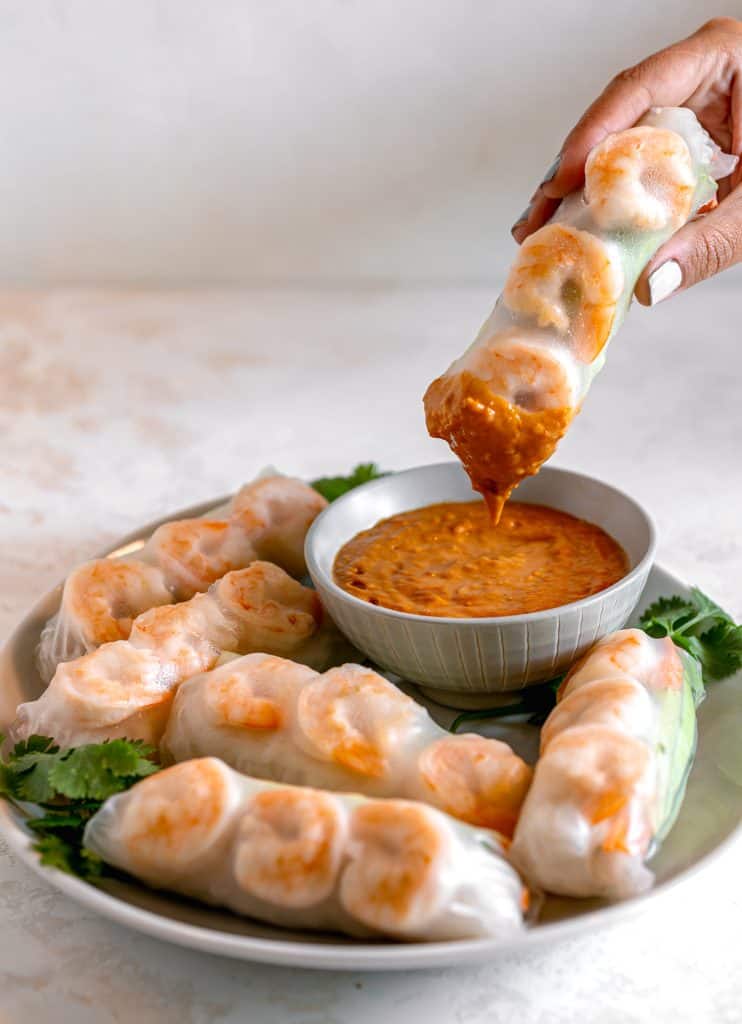 a shrimp spring roll being dipped into a bowl of spicy peanut sauce