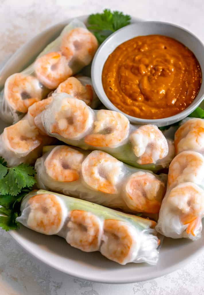 Shrimp spring rolls wrapped in rice paper on a white plate with a bowl of spicy peanut sauce in the right corner