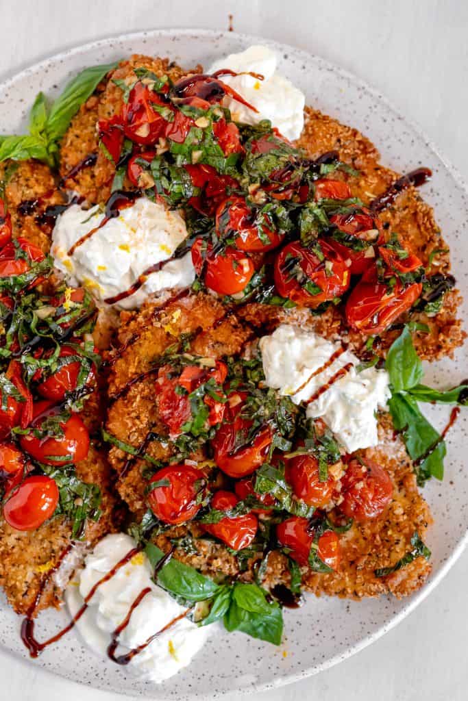 white spotted plate topped with crispy breaded chicken, tomato and basil salad, ripped burrata placed in between the chicken, and balsamic drizzle over the top on a white background