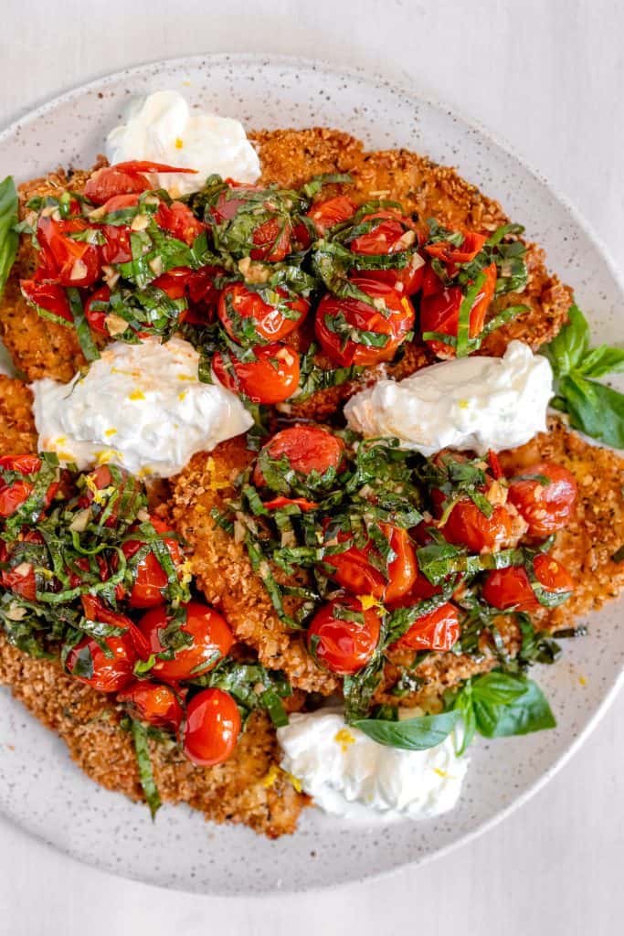white spotted plate topped with crispy breaded chicken, tomato and basil salad, ripped burrata placed in between the chicken, on a white background