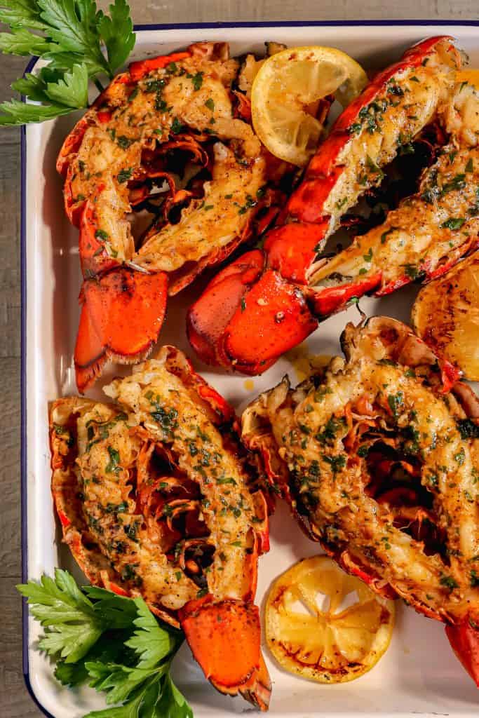 Four split open grilled lobster tails topped with garlic herb butter, surrounded by fresh parsley leaves, a grilled lemon, on a white pan
