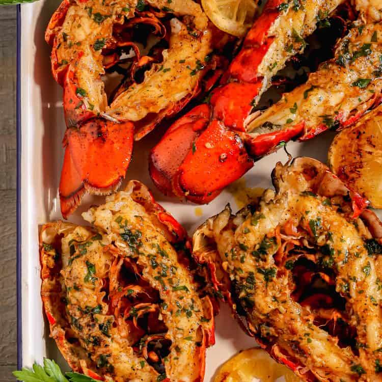 Four split open grilled lobster tails topped with garlic herb butter, surrounded by fresh parsley leaves, a grilled lemon, on a white pan